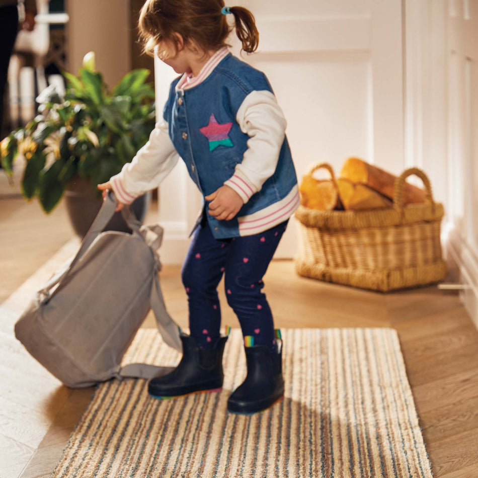 Child in rain boots standing on a rug with a suitcase indoors