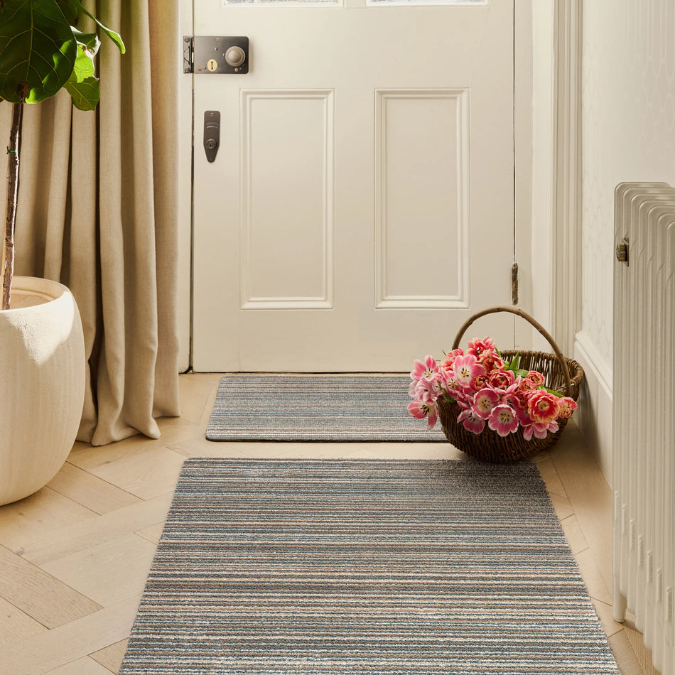 Front door with a basket of flowers on a striped rug, plant to the side.