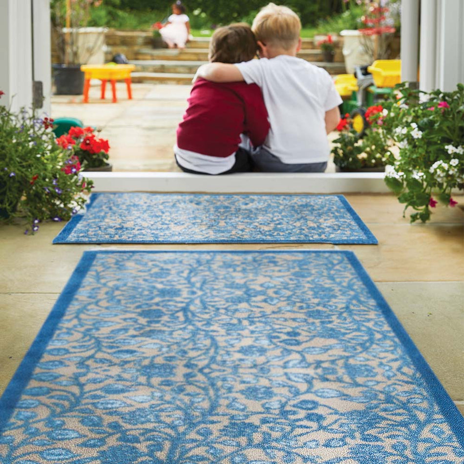 Two children sitting on a blue patterned rug in a garden setting with flowers and plants around. showing runner and mat