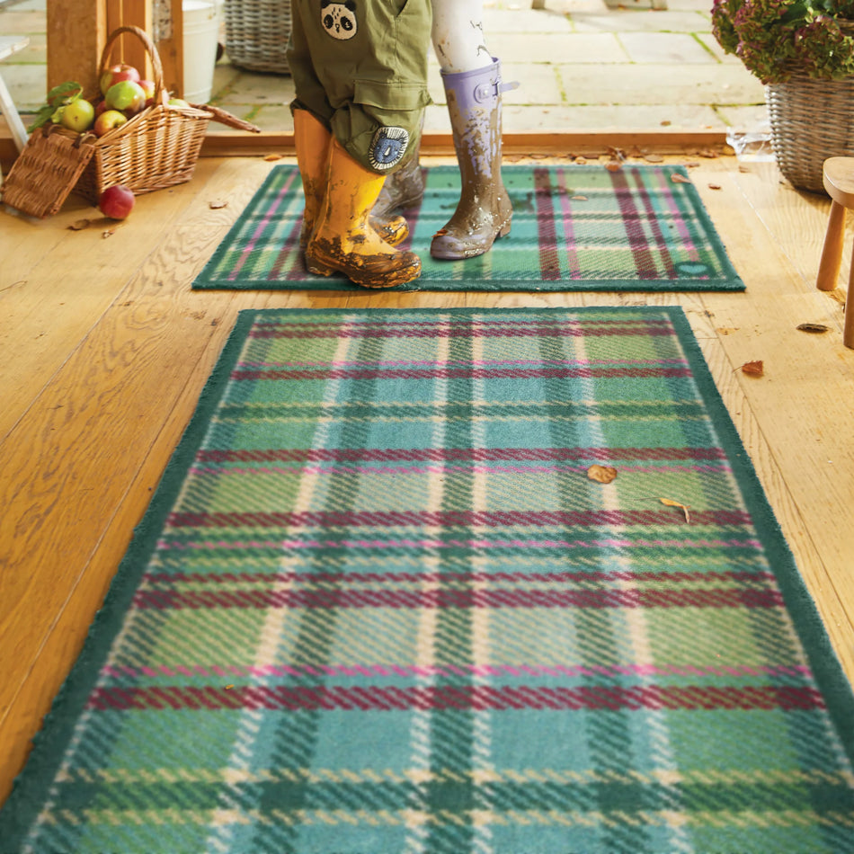Person wearing green wellies stepping onto a plaid rug on a wooden floor.