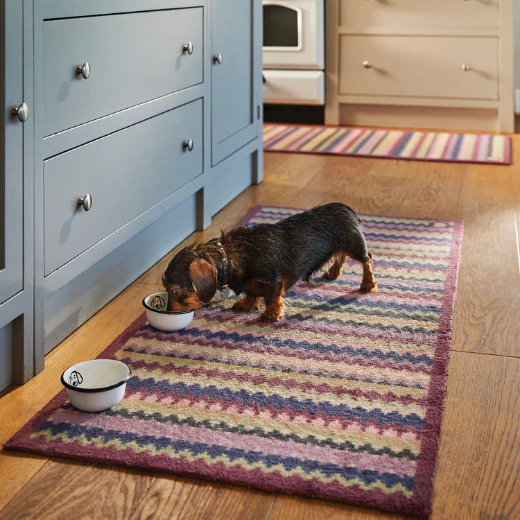 Small dog eating from a bowl on a colorful rug in a kitchen.