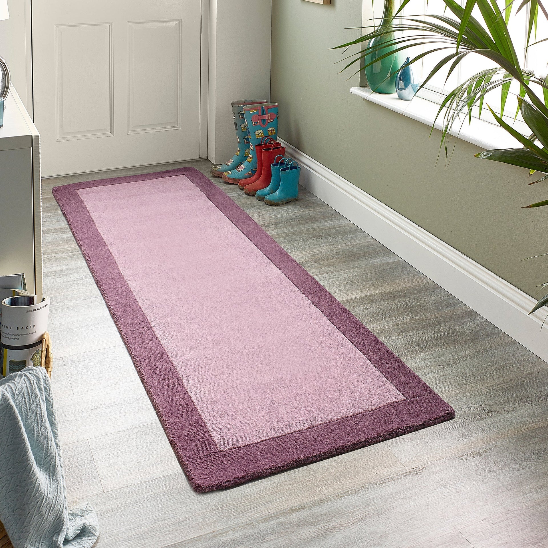Pink runner rug on a wooden floor with colorful boots and a plant in the background.