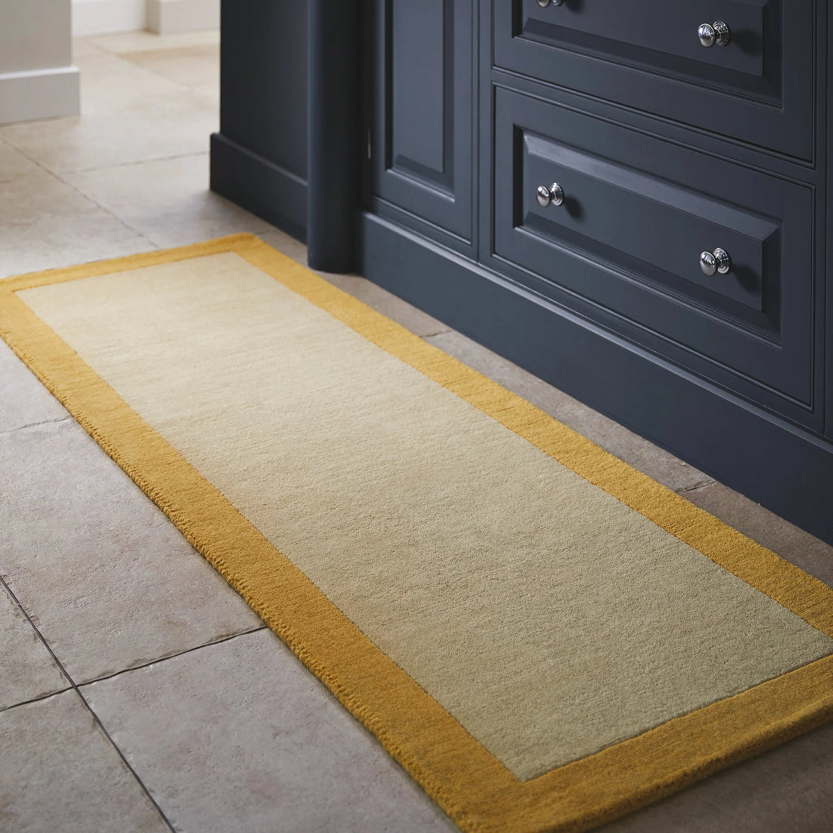 Beige rug with yellow border on a tiled floor next to dark blue cabinets.