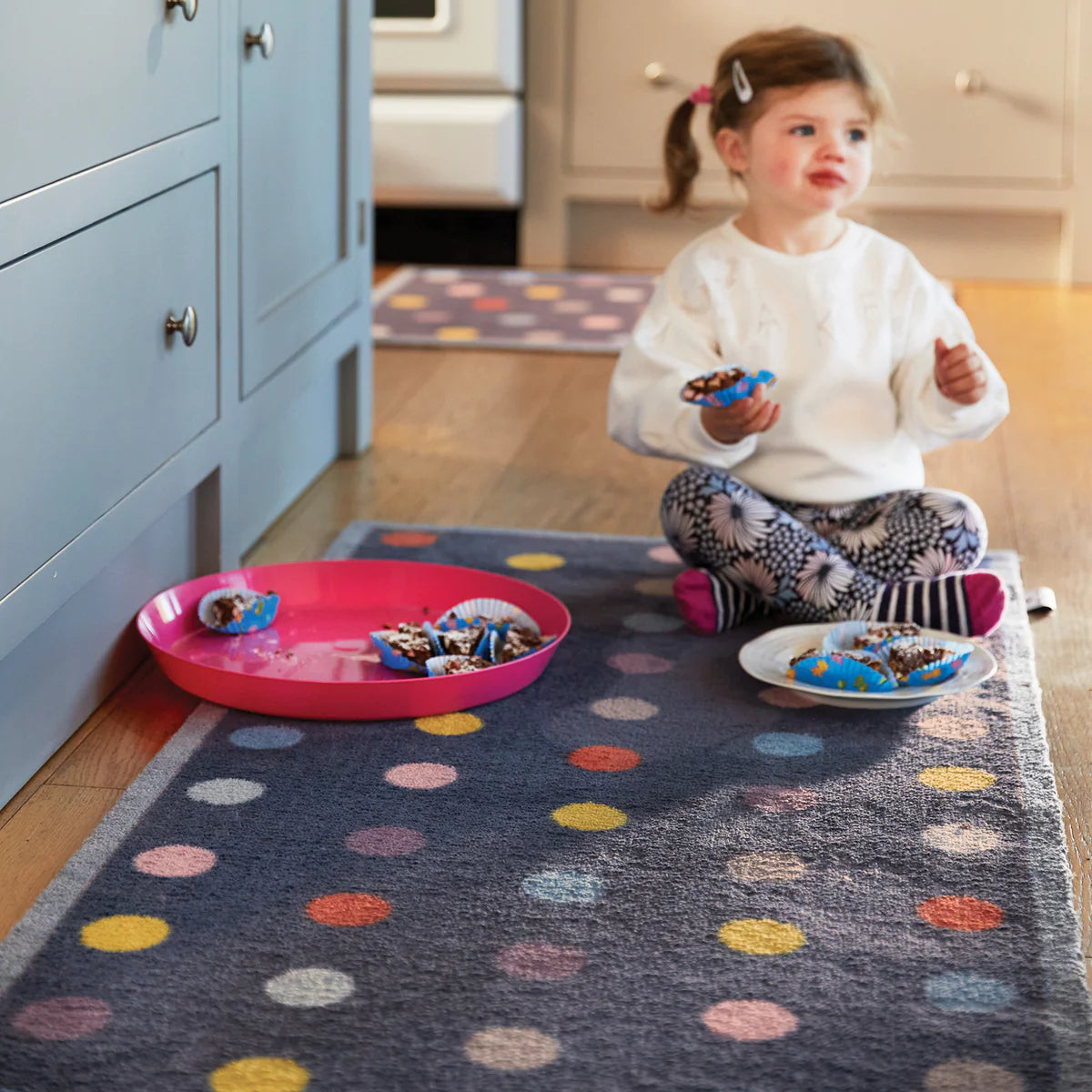 Child sitting on a colorful polka dot rug in a kitchen, eating snacks from plates.
