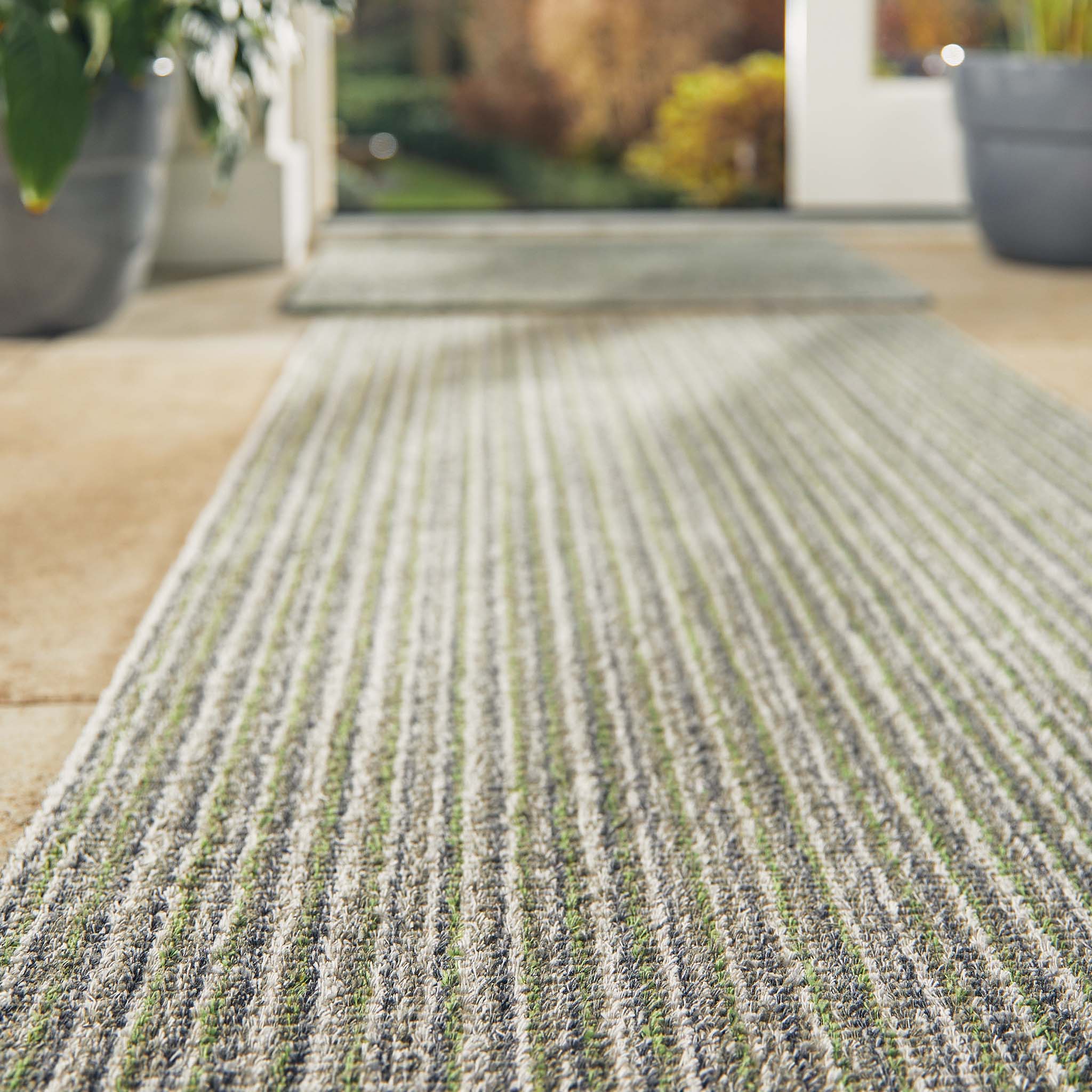 Close-up of a textured green and gray rug with potted plants in the background.
