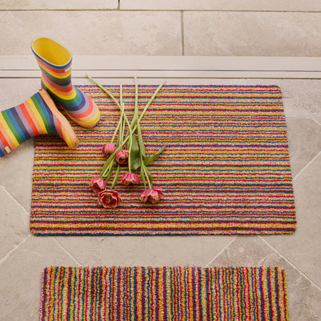 Colorful striped doormat with flowers and a rainbow boot on a tiled floor.
