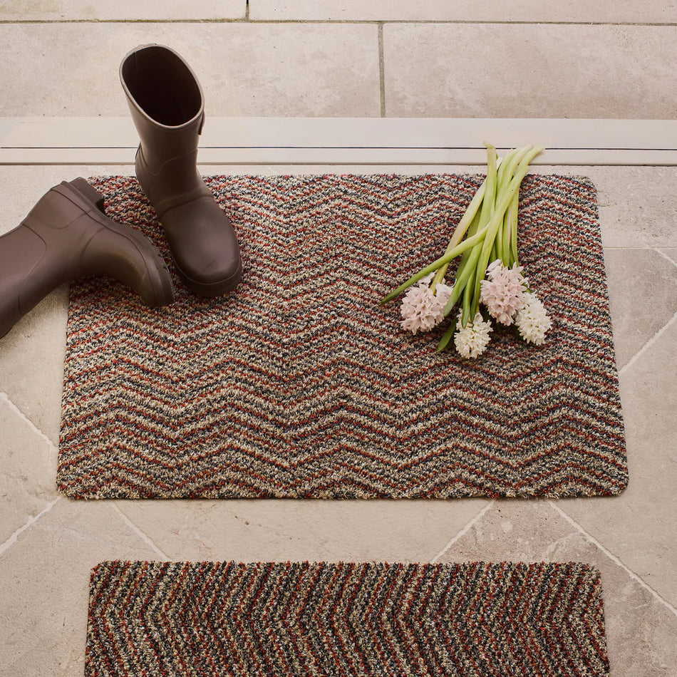 Doormat with zigzag pattern, rain boots, and flowers on a tiled floor.