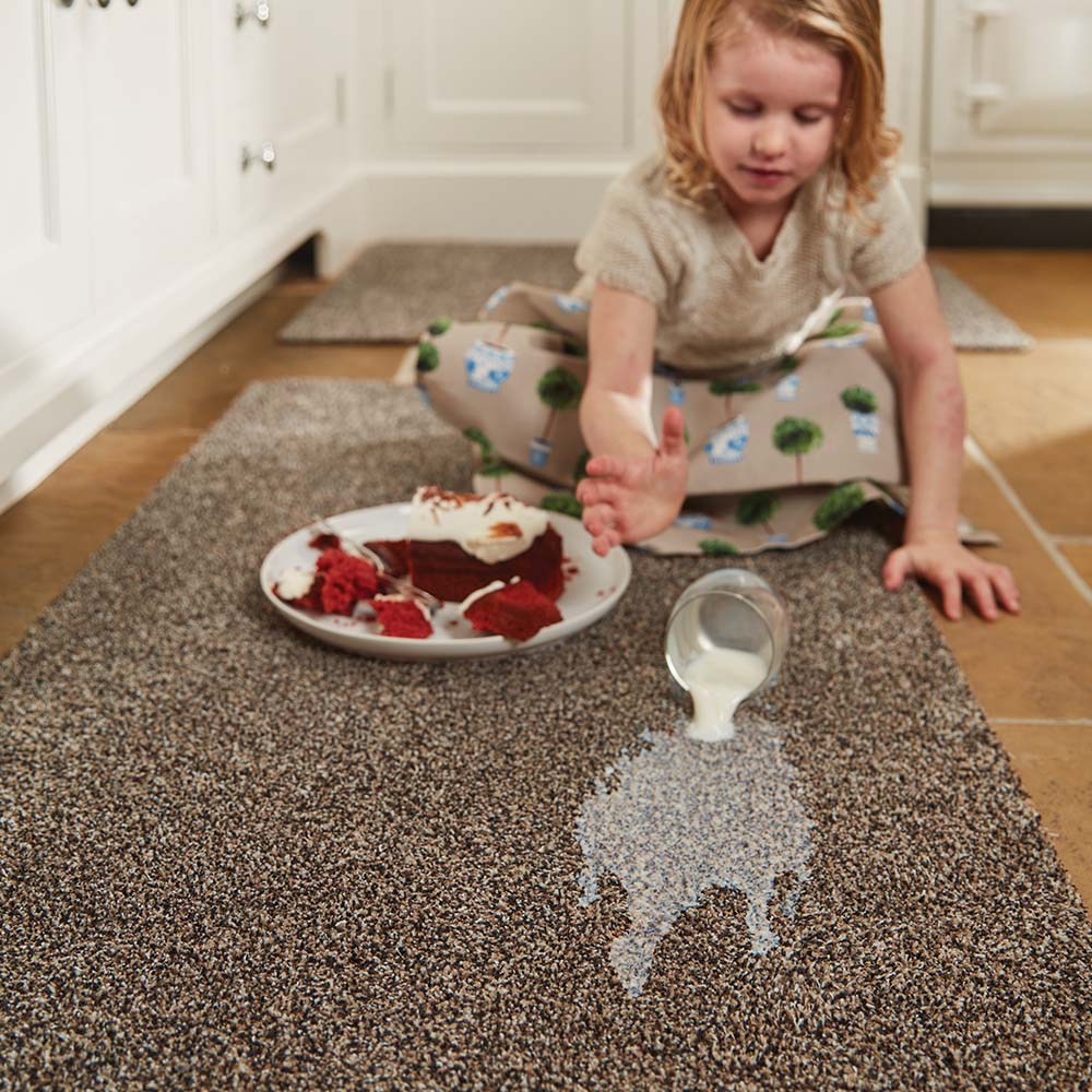 Child in a kitchen with a plate of cake and spilled milk on a carpeted floor.