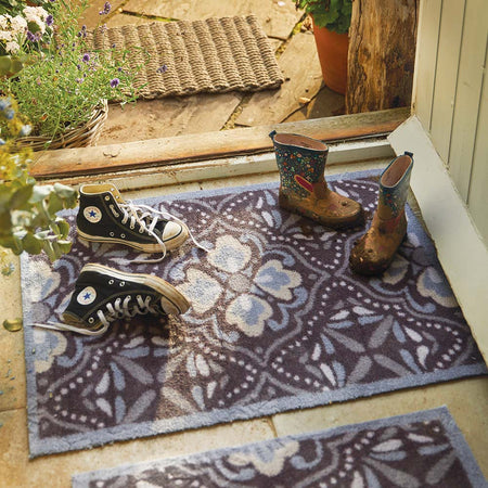 Two pairs of shoes on a decorative doormat outside a door with plants around.