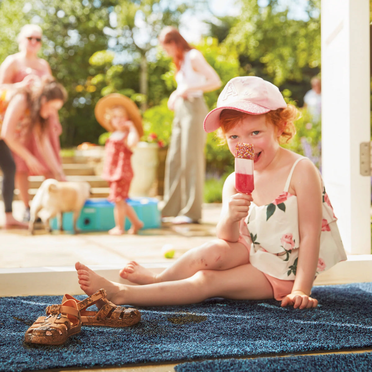 Child sitting on a blue rug outdoors, holding a popsicle with a family in the background.