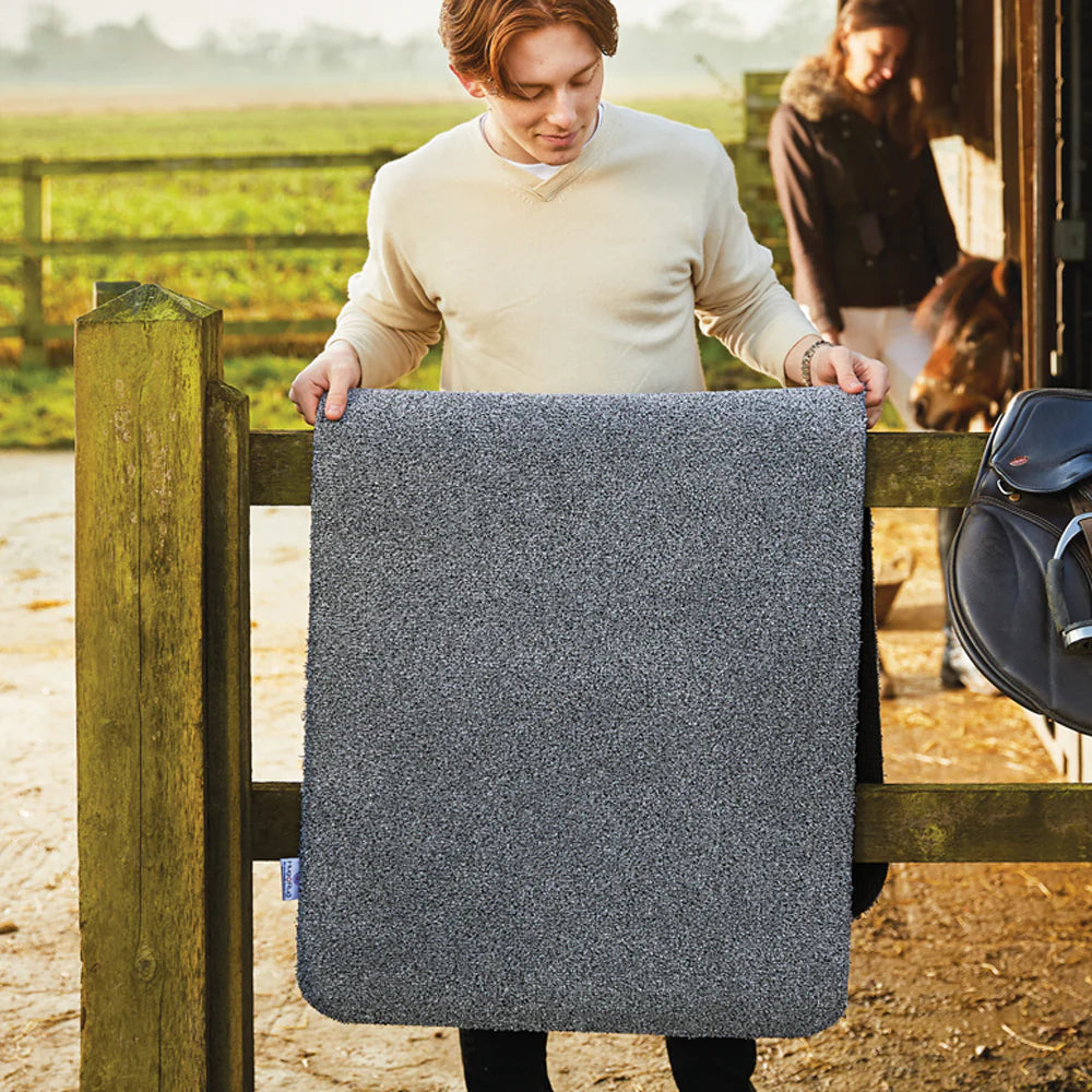Person holding a gray horse blanket in an outdoor setting with a horse and another person in the background.