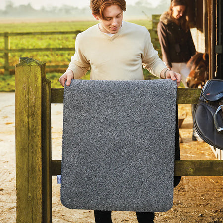 Person holding a gray horse blanket in an outdoor setting with a horse and another person in the background.