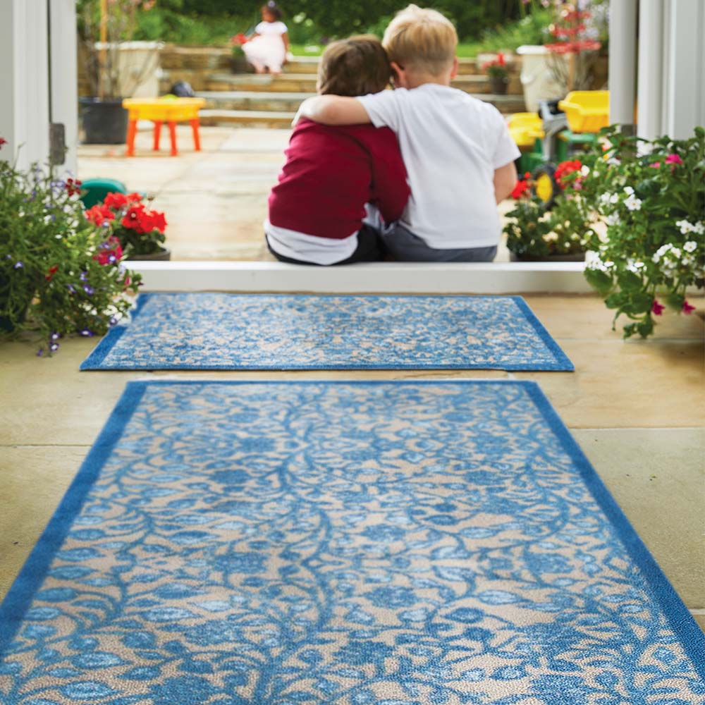 Two children sitting on a blue patterned rug in a garden setting with flowers and plants around. showing runner and mat