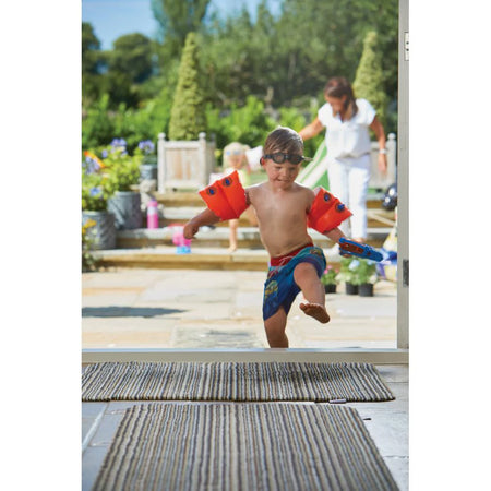 Child with red arm floats and blue swim trunks standing by a pool area.