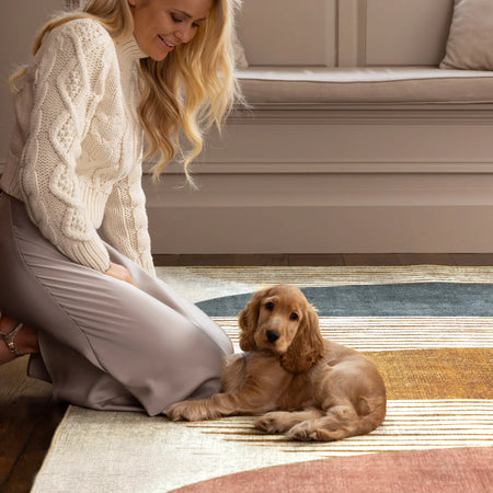 Woman in a white sweater and gray skirt kneeling on a striped rug with a small brown dog.