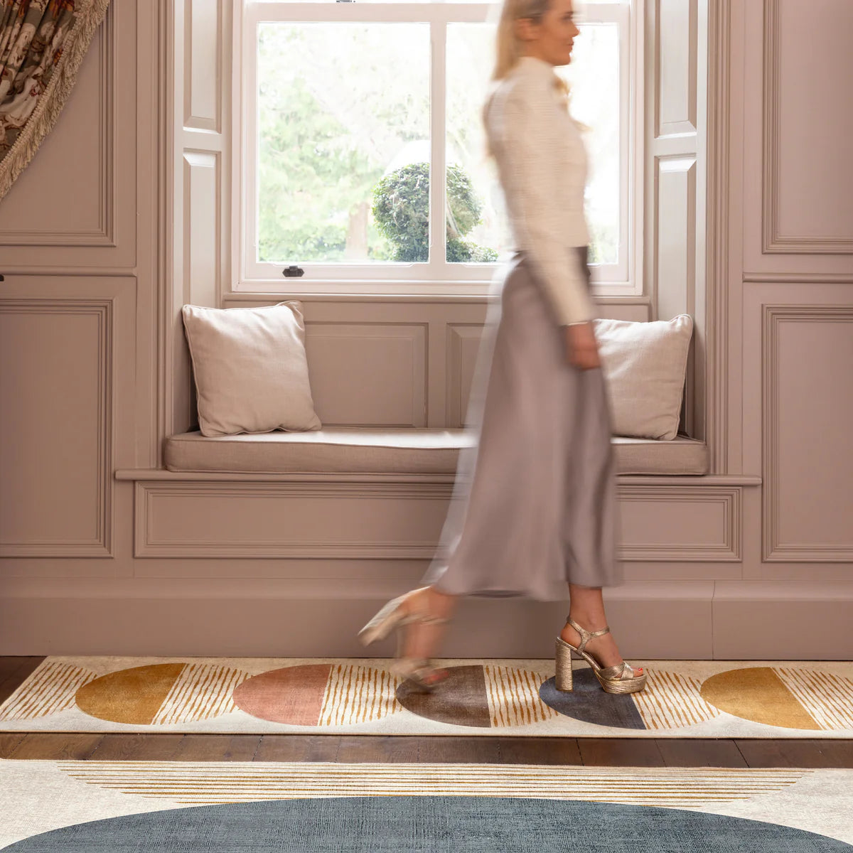 Woman walking past a window seat with cushions in a room with decorative wall paneling.