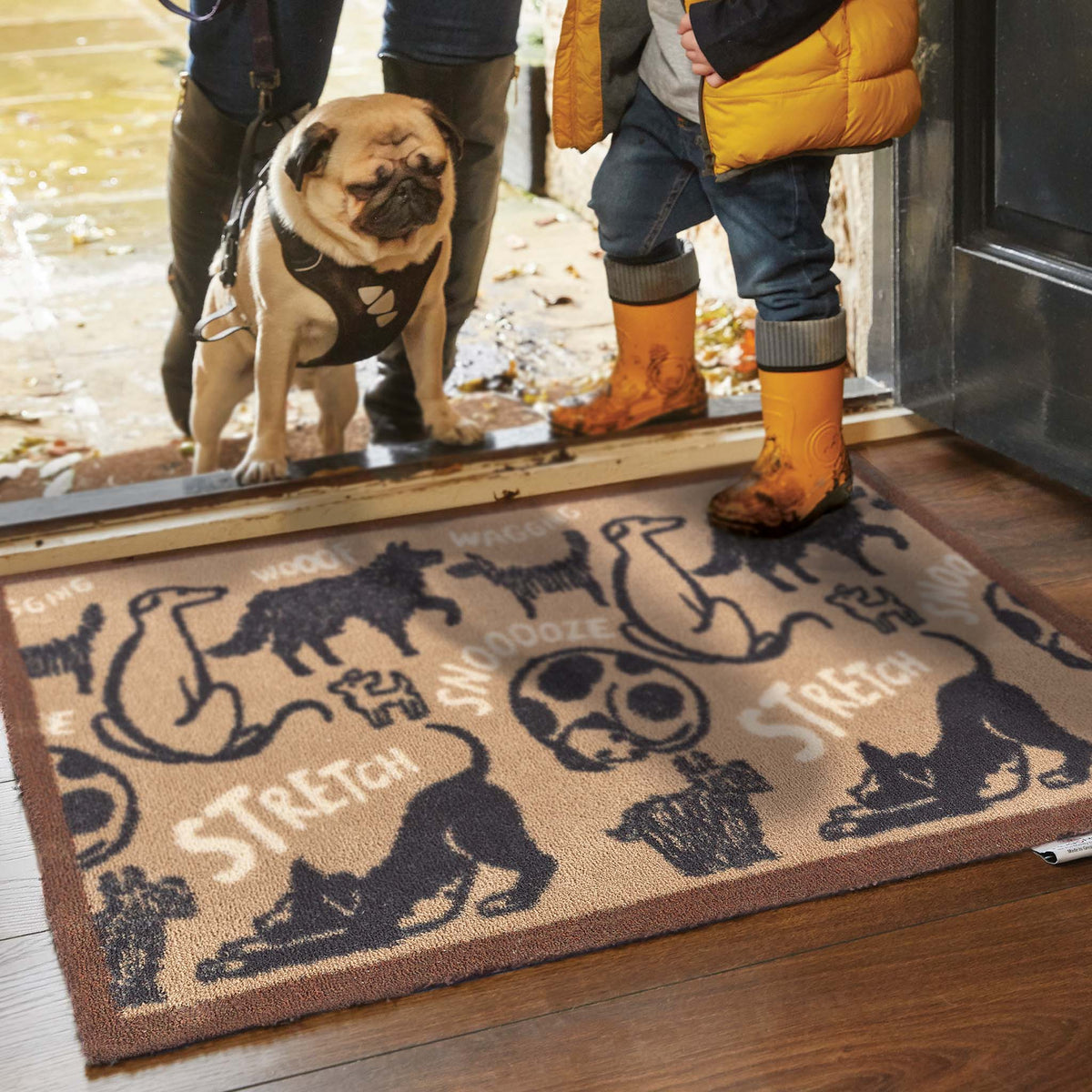 Dog stepping onto a doormat with animal illustrations and text, with a person in yellow boots nearby.