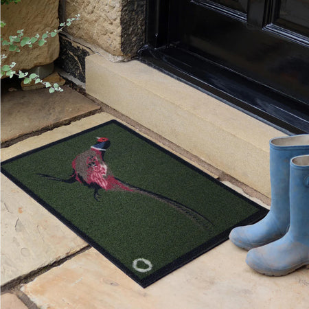 Doormat with a pheasant design on a stone floor next to a door, with blue boots nearby.