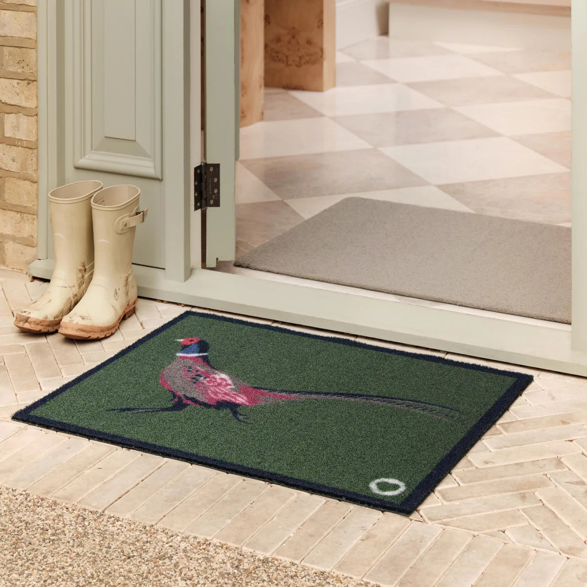 Green doormat with a pheasant design in front of a door with boots nearby.