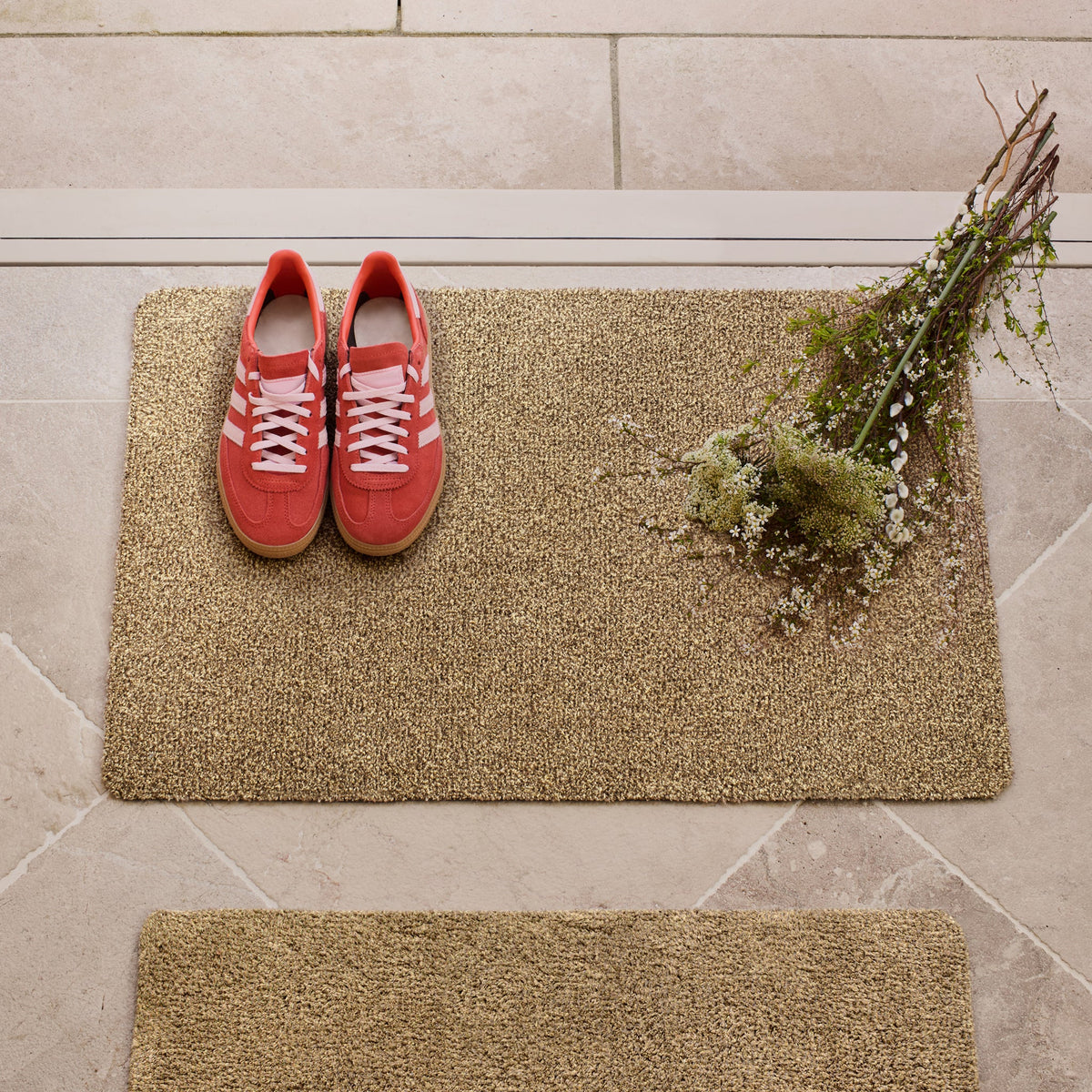Red trainers on a beige doormat with a plant on a stone floor.