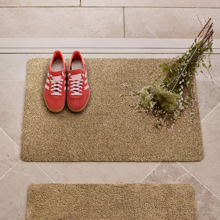 Red trainers on a beige doormat with a plant on a stone floor.