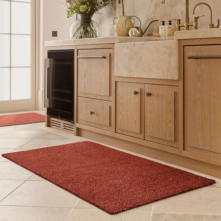 Red mat on a tiled floor in front of wooden kitchen cabinets with a window in the background.