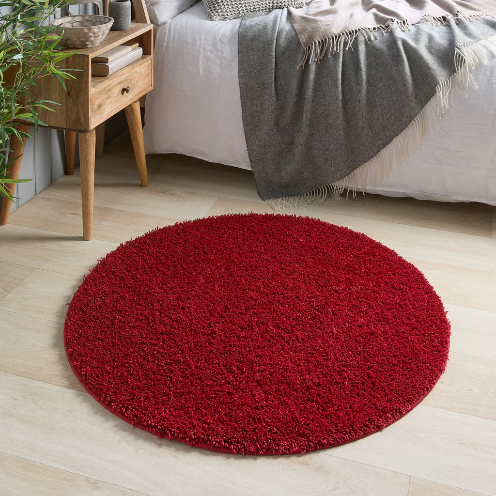Red round rug on a light wooden floor with a bed and side table in the background.