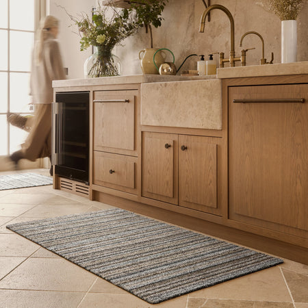 Kitchen with wooden cabinets, a rug on the floor, and a person walking by.