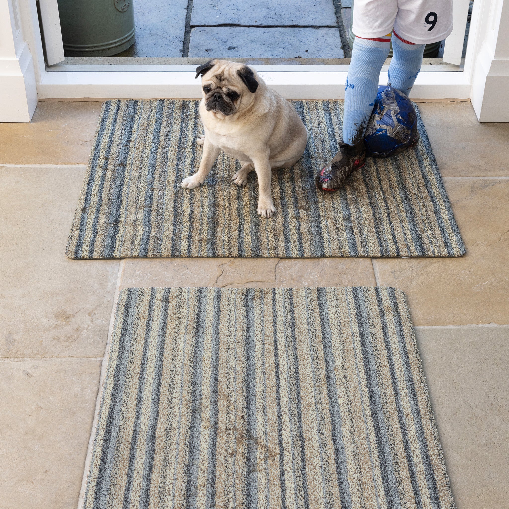 Pug sitting on a striped doormat with a person stepping onto another doormat.