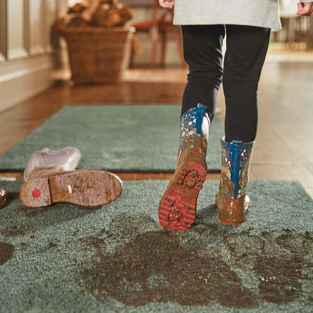 Person wearing colorful rain boots stepping onto a green rug with a blurred indoor background.