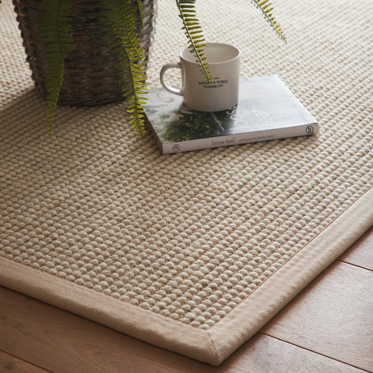 Sisal Boucle Beige woven mat on a wooden floor with a mug and book on top