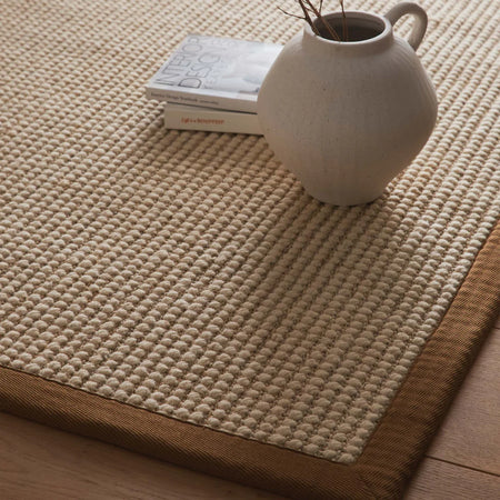 Beige woven mat with a white vase and books on top, on a wooden floor.