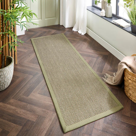 Long green woven Origins Sisal Sage Green with Sage Green Border
 rug on a wooden floor with plants and a basket in the background
