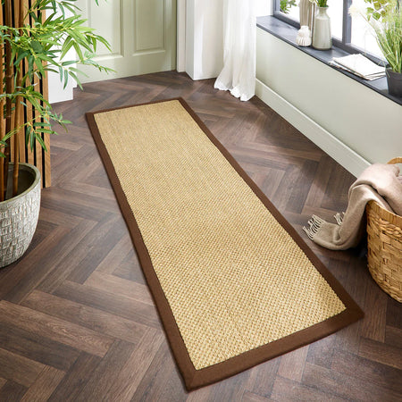Long woven sisal runner with border on a wooden floor with a plant and basket in the background